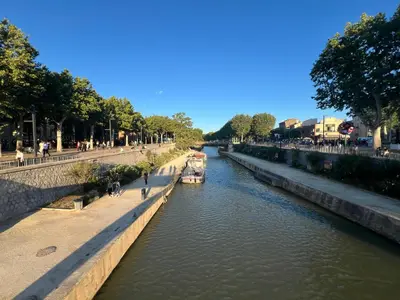a boat on a river with people walking on the sidewalk at La Villa des Cistes au calme vue Étangs in Narbonne