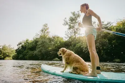 a woman standing on a paddle board with a dog at Camping Le Val de Cesse in Mirepeisset