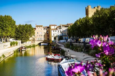 a river with boats and buildings and purple flowers at Studio Corneille -Climatisation WIFI Vue in Narbonne