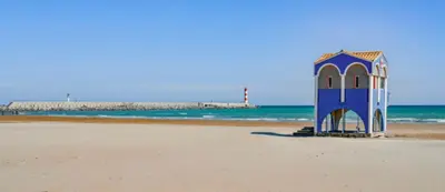 a blue building on the beach with a lighthouse in the background at La maison de Jade Le Mabrouka in Port-la-Nouvelle