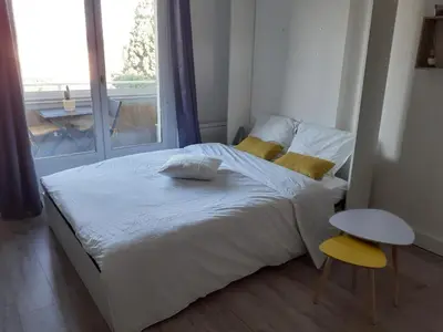 a bedroom with a white bed with a yellow stool and a window at Studio Balcon Proche Hypercentre de Narbonne in Narbonne