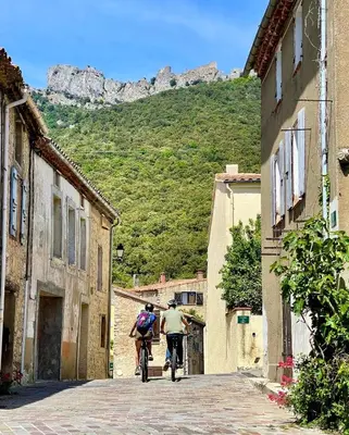 two people riding bikes down a street with mountains in the background at Gîte XL Familles et groupes Duilhac sous Peyrepertuse in Duilhac